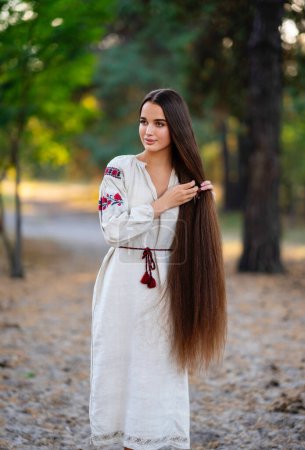 Téléchargez les photos : Belle fille cheveux tressés extra longue robe traditionnelle ukrainienne posant dans la forêt. Portrait de jeune femme élégante attrayante sur fond chaud coloré. - en image libre de droit
