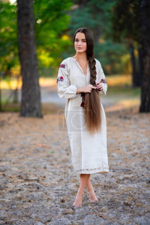 Photo for Beautiful extra long braided hair girl in Ukrainian traditional dress posing in forest . Portrait of young attractive stylish woman on colorful warm background. - Royalty Free Image