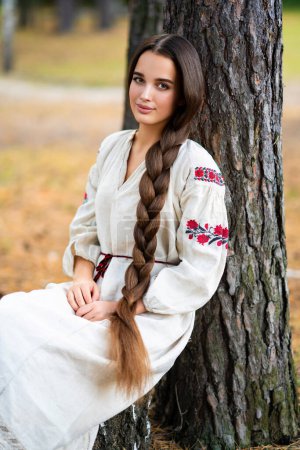 Téléchargez les photos : Belle fille cheveux tressés extra longue robe traditionnelle ukrainienne posant dans la forêt. Portrait de jeune femme élégante attrayante sur fond chaud coloré. - en image libre de droit
