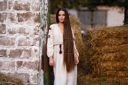 Téléchargez les photos : Belle fille de cheveux tressés extra longue en robe traditionnelle ukrainienne posant sur une ferme. Portrait de jeune femme élégante attrayante sur fond chaud coloré. - en image libre de droit
