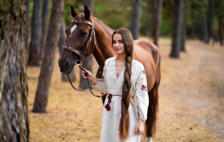 Beautiful extra long braided hair girl in Ukrainian traditional dress posing with horse in forest . Portrait of young attractive stylish woman on colorful warm background.