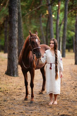 Téléchargez les photos : Belle fille cheveux tressés extra longue robe traditionnelle ukrainienne posant avec cheval dans la forêt. Portrait de jeune femme élégante attrayante sur fond chaud coloré. - en image libre de droit