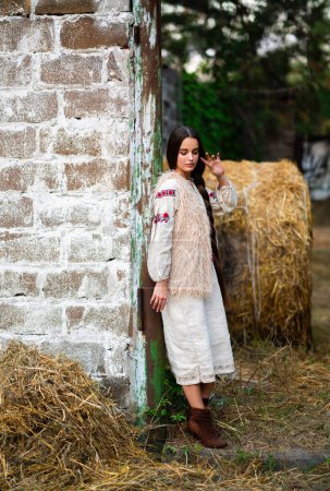 Téléchargez les photos : Belle fille de cheveux tressés extra longue en robe traditionnelle ukrainienne posant sur une ferme. Portrait de jeune femme élégante attrayante sur fond chaud coloré. - en image libre de droit