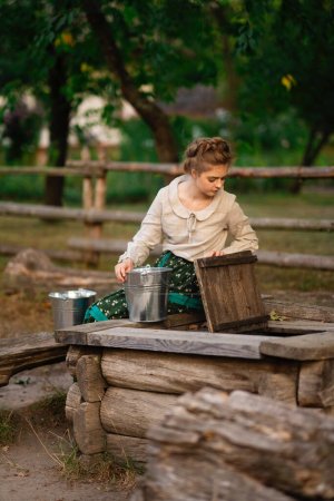 Foto de Hermosa chica trenzada rubia en vestido largo rural posando en un pueblo con cubos de agua. Bastante joven modelo en campo arte foto. - Imagen libre de derechos