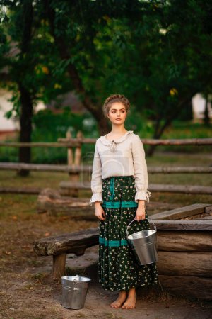 Foto de Hermosa chica trenzada rubia en vestido largo rural posando en un pueblo con cubos de agua. Bastante joven modelo en campo arte foto. - Imagen libre de derechos