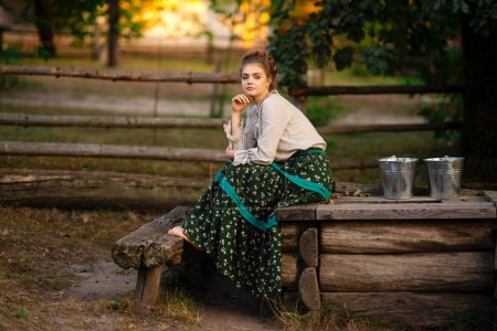 Photo for Beautiful  blonde braided girl in rural long dress posing in a village with buckets of water. Pretty young model in countryside art photo. - Royalty Free Image