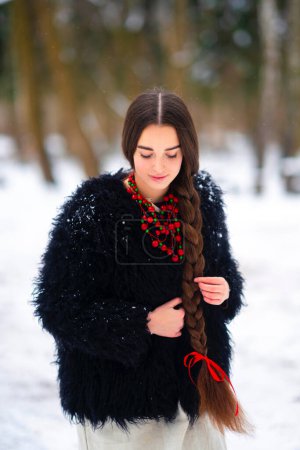 Photo for Beautiful extra long braided hair girl in Ukrainian traditional dress, black fur coat and red beads posing in winter forest . Portrait of young attractive stylish woman on winter background. - Royalty Free Image