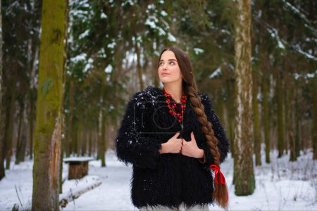 Photo for Beautiful extra long braided hair girl in Ukrainian traditional dress, black fur coat and red beads posing in winter forest . Portrait of young attractive stylish woman on winter background. - Royalty Free Image