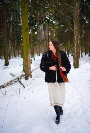 Téléchargez les photos : Belle fille très longue cheveux tressés en robe traditionnelle ukrainienne, manteau en fourrure noire et perles rouges posant dans la forêt d'hiver. Portrait de jeune femme élégante attrayante sur fond d'hiver. - en image libre de droit