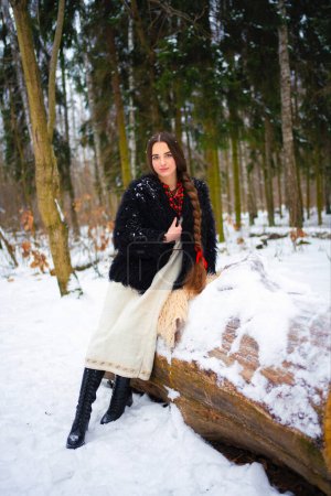 Téléchargez les photos : Belle fille très longue cheveux tressés en robe traditionnelle ukrainienne, manteau en fourrure noire et perles rouges posant dans la forêt d'hiver. Portrait de jeune femme élégante attrayante sur fond d'hiver. - en image libre de droit