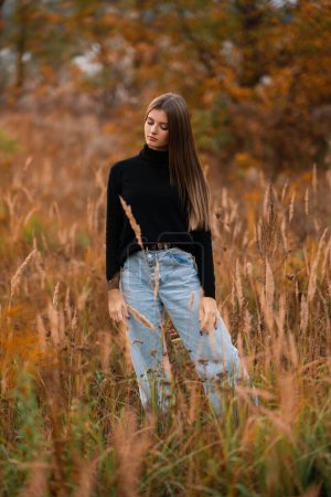 Téléchargez les photos : Belle fille portant un jean et un golf noir profitant de la nature d'automne en plein air. Jeune joli modèle posant sur fond orange. - en image libre de droit