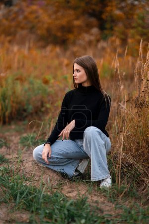 Foto de Hermosa chica con jeans y golf negro disfrutando de la naturaleza otoñal al aire libre. Joven modelo bonita posando sobre fondo naranja. - Imagen libre de derechos