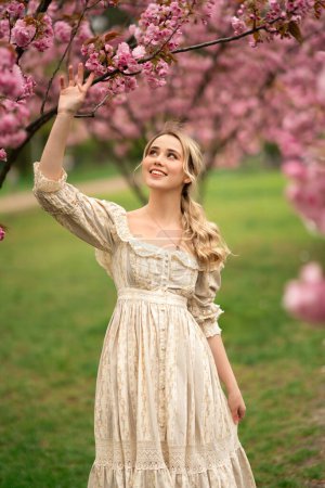 Téléchargez les photos : Jolie jeune fille blonde en robe de dentelle vintage debout dans le parc de printemps près de fleurs roses. Tendresse modèle romantique posant. - en image libre de droit
