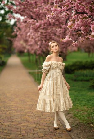 Foto de Chica rubia bastante joven en vestido de encaje vintage de pie en el parque de primavera cerca de flores de flor rosa. Ternura romántico modelo posando. - Imagen libre de derechos