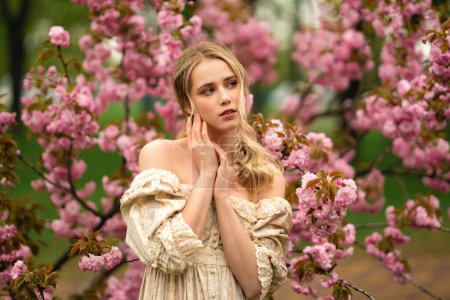 Téléchargez les photos : Jolie jeune fille blonde en robe de dentelle vintage debout dans le parc de printemps près de fleurs roses. Tendresse modèle romantique posant. - en image libre de droit