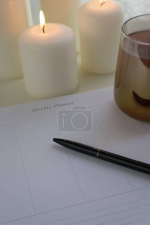 Photo for A black pen lies on empty sheets of a weekly planner next to a cup of tea and candles copy space - Royalty Free Image