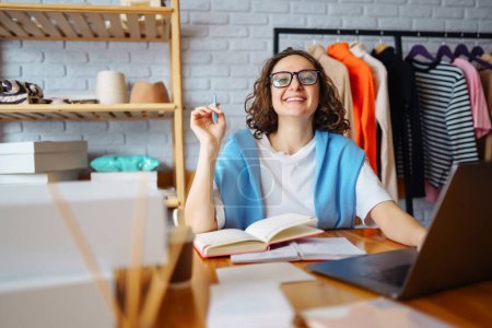 Photo for Young female dress designer sitting at workplace with laptop. Small business owner, entrepreneur, seller checking ecommerce clothing store orders. Shopping concept. Fashion studio. - Royalty Free Image