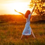 Young woman in stylish summer dress feeling free in the field with flowers at sunset. Nature, fashion, vacation and lifestyle