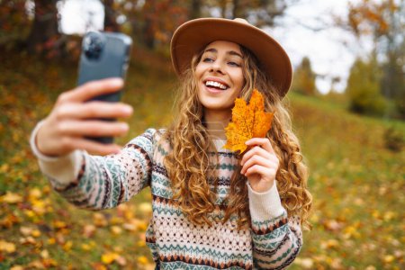 Beautiful woman in a stylish sweater with a phone in her hands sits in a clearing among yellow fallen leaves in an autumn park. Tourist enjoys the weather, takes a photo with a yellow leaf. 