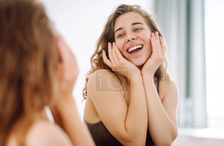 Téléchargez les photos : Une jeune femme regarde dans le miroir examiner l'inflammation sur son visage. Peau d'acné. Espace de copie. Médecine et cosmétologie. - en image libre de droit