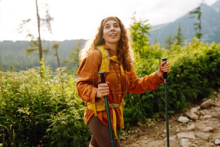Young woman traveler with a yellow hiking backpack and hiking poles on trail among the mountains. Adventure concept, hiking. Active lifestyle.