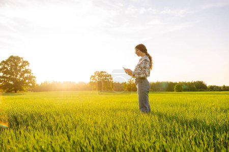 Photo for Young woman agronomist works on a modern digital tablet on a green wheat field. A senior woman farmer checks the quality of young wheat sprouts using a tablet. - Royalty Free Image