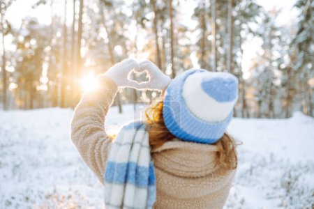 Una mujer con un sombrero y una bufanda contra el telón de fondo de un bosque invernal nevado sostiene sus manos en forma de corazón. Vista trasera. Mujer joven disfrutando de un día soleado de invierno. Concepto de vacaciones, estilo de vida.