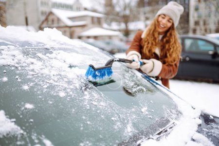 Nettoyage de la neige d'une voiture. Une belle femme nettoie la neige d'une voiture avec une brosse. Concept de transport, saisonnalité Nettoyage hivernal du verre.
