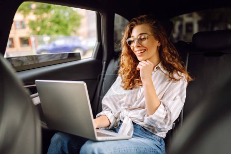 Photo for Young woman with laptop while sitting in the back seat of a car. Education online. Remote work. Business, blogging, freelance concept. - Royalty Free Image