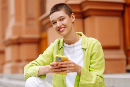 Joyful beautiful woman using cellphone while sitting on stairs outdoors. Using smartphone apps. Concept of technology, tourism, urban life.