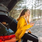 Young woman sitting in open trunk with phone. Lifestyle, travel, tourism, nature, active life.