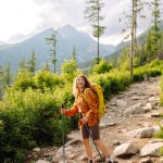 Backpacker in a hike in the summer mountains. Young woman with a yellow backpack and hiking equipment walks along a hiking trail against the backdrop of mountain scenery.