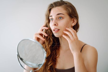 Mujer joven con manchas rojas infeliz por el acné y las imperfecciones de la piel, mirando a la reflexión en el espejo en el baño. Cuidado facial de la piel. Problema del acné.