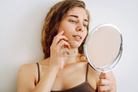 Jeune femme avec tache rouge malheureux sur l'acné et les imperfections de la peau, en regardant le reflet dans le miroir dans la salle de bain. Soins du visage. Problème d'acné.