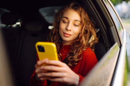 Téléchargez les photos : Portrait d'une femme bouclée avec un smartphone dans les mains sur le siège arrière d'une voiture. Concept de technologie, voyage en voiture, affaires. - en image libre de droit