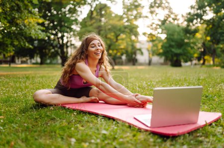 Téléchargez les photos : Jeune femme suit avec un ordinateur portable un exercice d'ajustement dans le parc. Formation en ligne, cours. Entraînement. Mode de vie sain, sport. - en image libre de droit