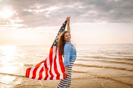 Téléchargez les photos : Femme patriote avec drapeau américain sur la plage au coucher du soleil. Les USA célèbrent le 4 juillet. Concept de fête de l'indépendance - en image libre de droit