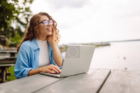 Beautiful woman sitting on a bench at a table in a park outdoors, using a laptop. Concept for education, business, blog or freelance.
