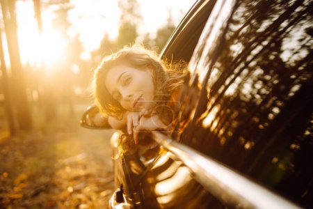 Téléchargez les photos : Jeune femme assise en voiture et penchée sur la fenêtre pendant son road trip et profitant d'une belle journée dans la nature. Mode de vie, week-end, tourisme, nature, vie active. - en image libre de droit
