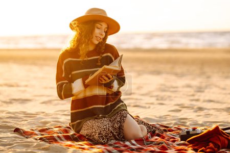 Téléchargez les photos : Jeune femme dans un pull confortable et chapeau aime la nature, la lecture d'un livre tout en étant assis sur une couverture au pique-nique de la plage. Le concept de détente, de plaisir, de solitude avec la nature. - en image libre de droit