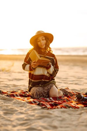 Téléchargez les photos : Jeune femme dans un pull confortable et chapeau aime la nature, la lecture d'un livre tout en étant assis sur une couverture au pique-nique de la plage. Le concept de détente, de plaisir, de solitude avec la nature. - en image libre de droit