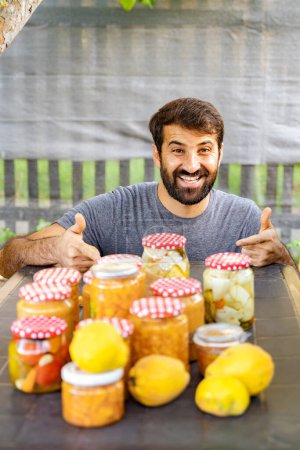 Happy eastern indian pakistani man is smiling, giving thumbs up near table full of conserves, jams and jars and lemons, producing natural products from his garden. Hobby or small business