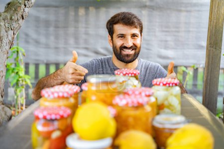 Photo for Happy eastern indian pakistani man is smiling, giving thumbs up near table full of conserves, jams and jars and lemons, producing natural products from his garden. Hobby or small business - Royalty Free Image