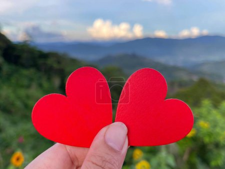 Photo for View of left hand holding two hearts shaped of red papers on blurred background of la green landscape. - Royalty Free Image