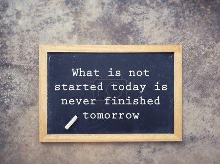 Photo for Motivational and inspirational wording. What Is Not Started Today Is Never Finished Tomorrow written on a blackboard. With blurred styled background. - Royalty Free Image