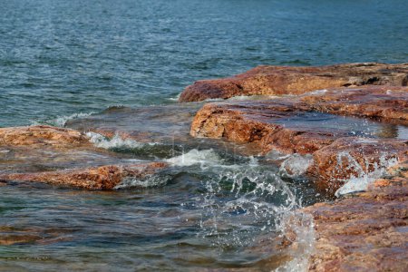 Lots of small waves hitting red and black beach rocks in Eira aka Eiranranta, Helsinki, Finland. Sunny summer day by the Baltic Sea. Color image showing the power of nature, the sea, and clean Nordic city nature.