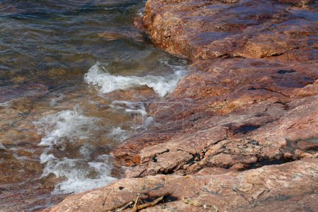 Lots of small waves hitting red and black beach rocks in Eira aka Eiranranta, Helsinki, Finland. Sunny summer day by the Baltic Sea. Color image showing the power of nature, the sea, and clean Nordic city nature.