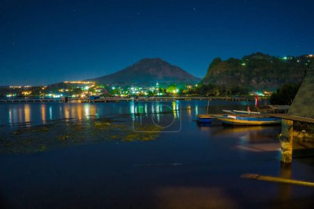 Die Abendstimmung am Lake Batur, Kintamani, Bali, mit der Reflexion des Mount Batur und dem Ambiente des Dorfes am Ufer des Sees, das von glitzernden Lichtern erleuchtet wird. Nachtfotografie.