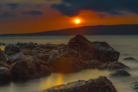 Photo for The stunning sunrise view in the Bali Strait, Indonesia, with a foreground of rocky shores along the beach, was captured using the long exposure photography technique. Landscape photography. - Royalty Free Image
