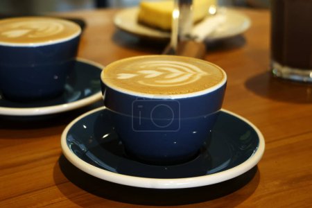 Foto de Una taza de cerámica llena de café se sienta en un platillo azul colocado en una mesa de madera. Delicado arte de latte blanco adorna la superficie del café. - Imagen libre de derechos